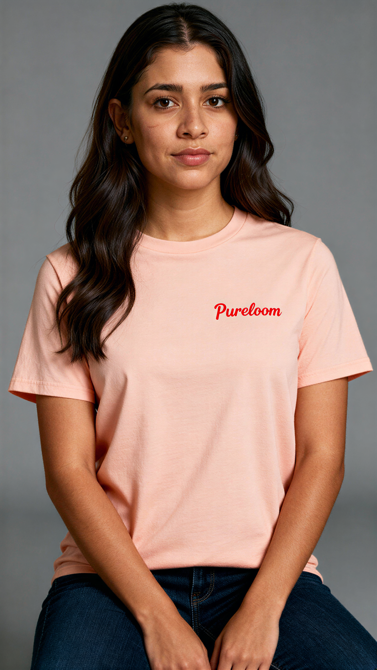 במבט קדמי, אישה לובשת חולצת כותנה אורגנית בצבע ורוד עם לוגו אדום "Pureloom" בצד שמאל של החזה מבית פיורלום.