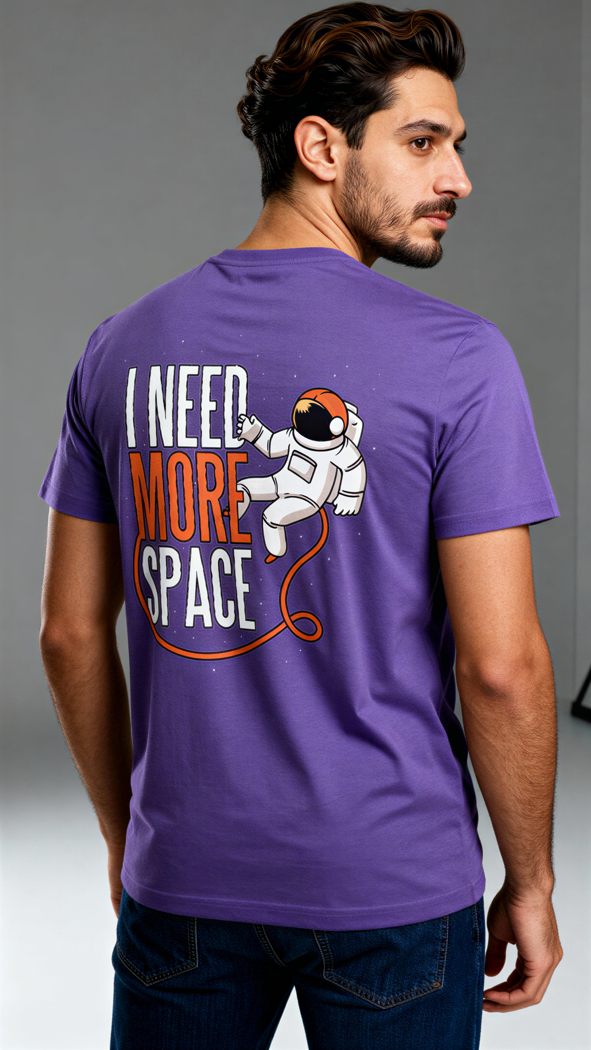 במבט מאחור, גבר לובש חולצת כותנה אורגנית סגולה עם הדפס של אסטרונאוט וכיתוב "I Need More Space" בעיצוב צבעוני מבית פיורלום.