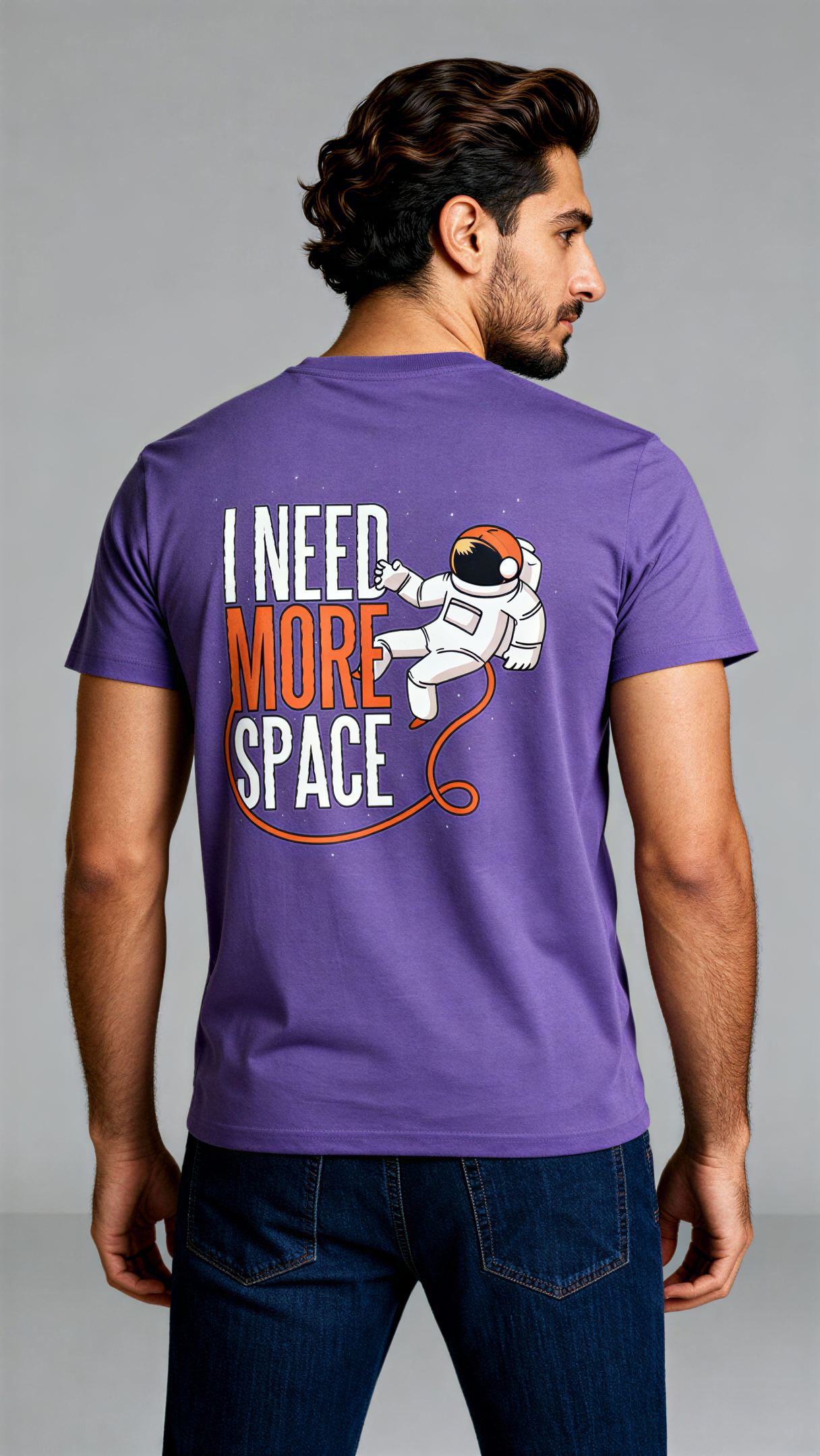 במבט מאחור, גבר לובש חולצת כותנה אורגנית סגולה עם הדפס של אסטרונאוט וכיתוב "I Need More Space" בעיצוב צבעוני מבית פיורלום.