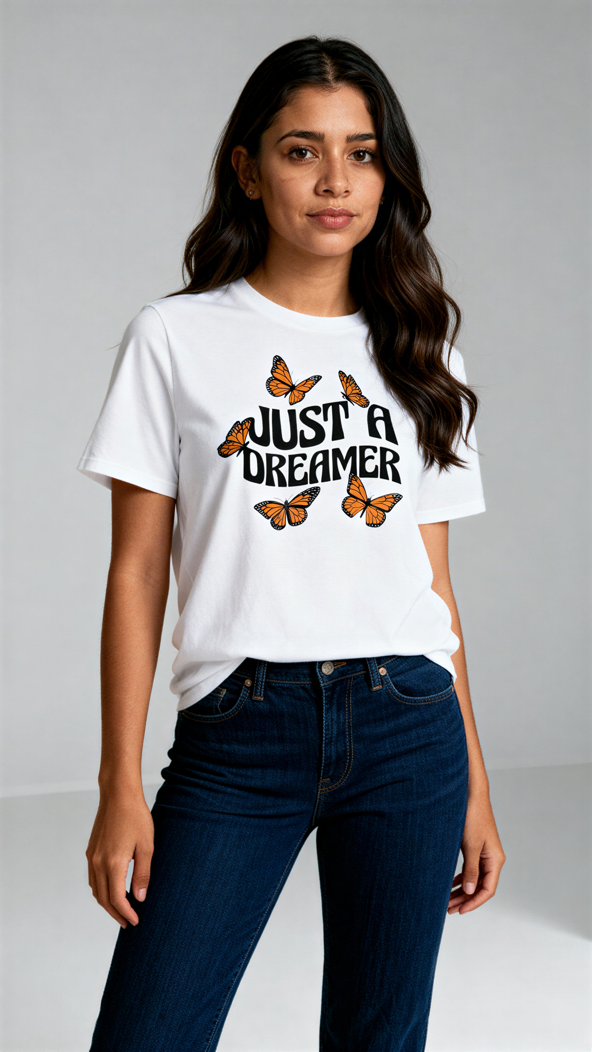 במבט קדמי, אישה לובשת חולצת כותנה אורגנית לבנה עם הדפס של פרפרים כתומים וכיתוב שחור גדול "Just a Dreamer" מבית פיורלום.