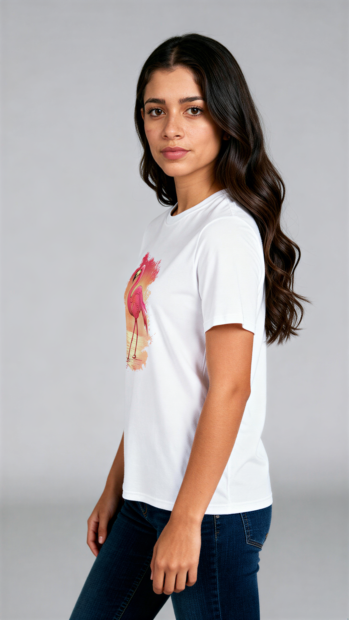במבט צדדי, אישה לובשת חולצת כותנה אורגנית לבנה עם הדפס צבעוני של פלמינגו עומד במים על רקע שקיעה בגווני כתום וורוד מבית פיורלום.